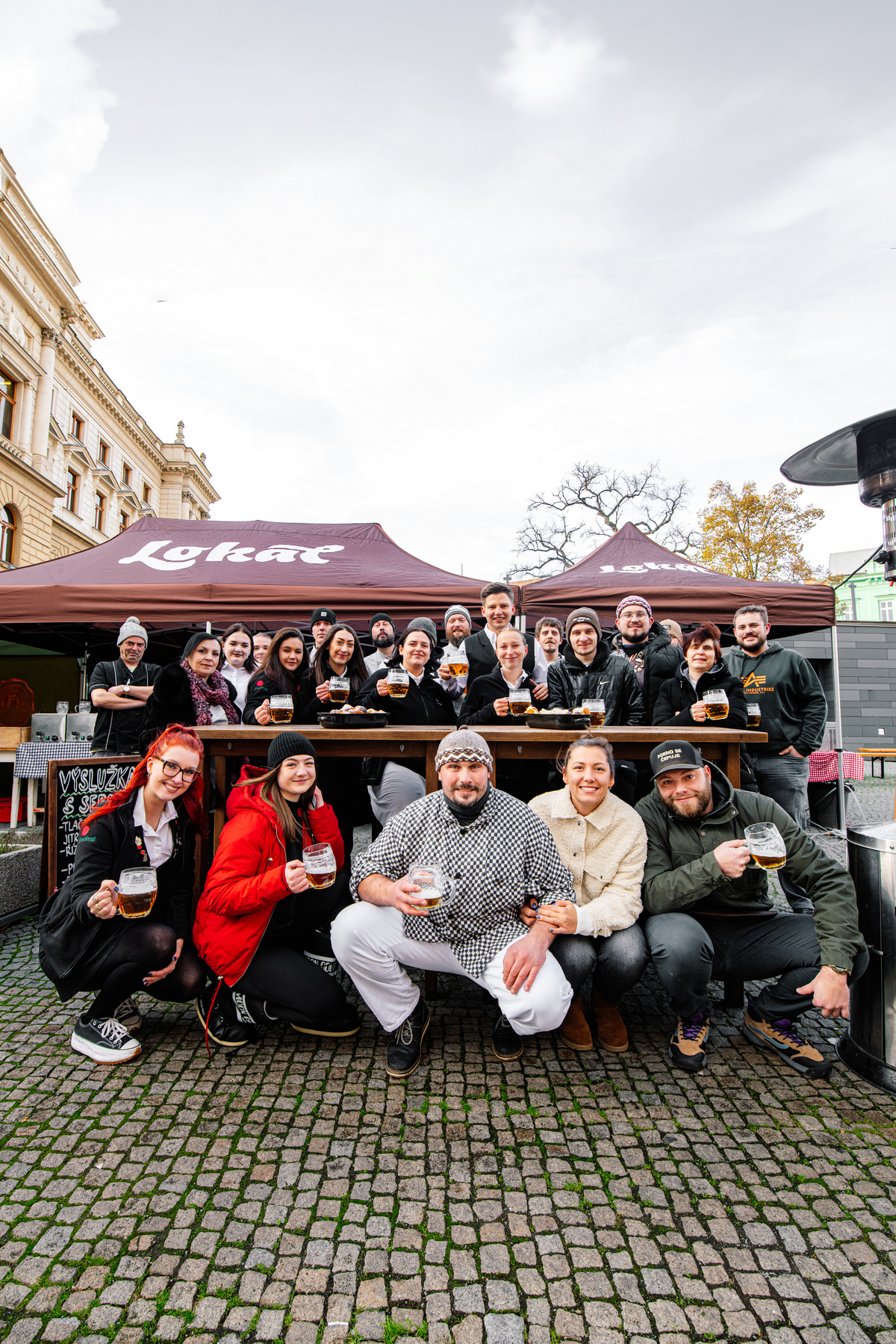 Skupina lidí pózuje s půllitry piva před stánky Lokálu na venkovní akci.