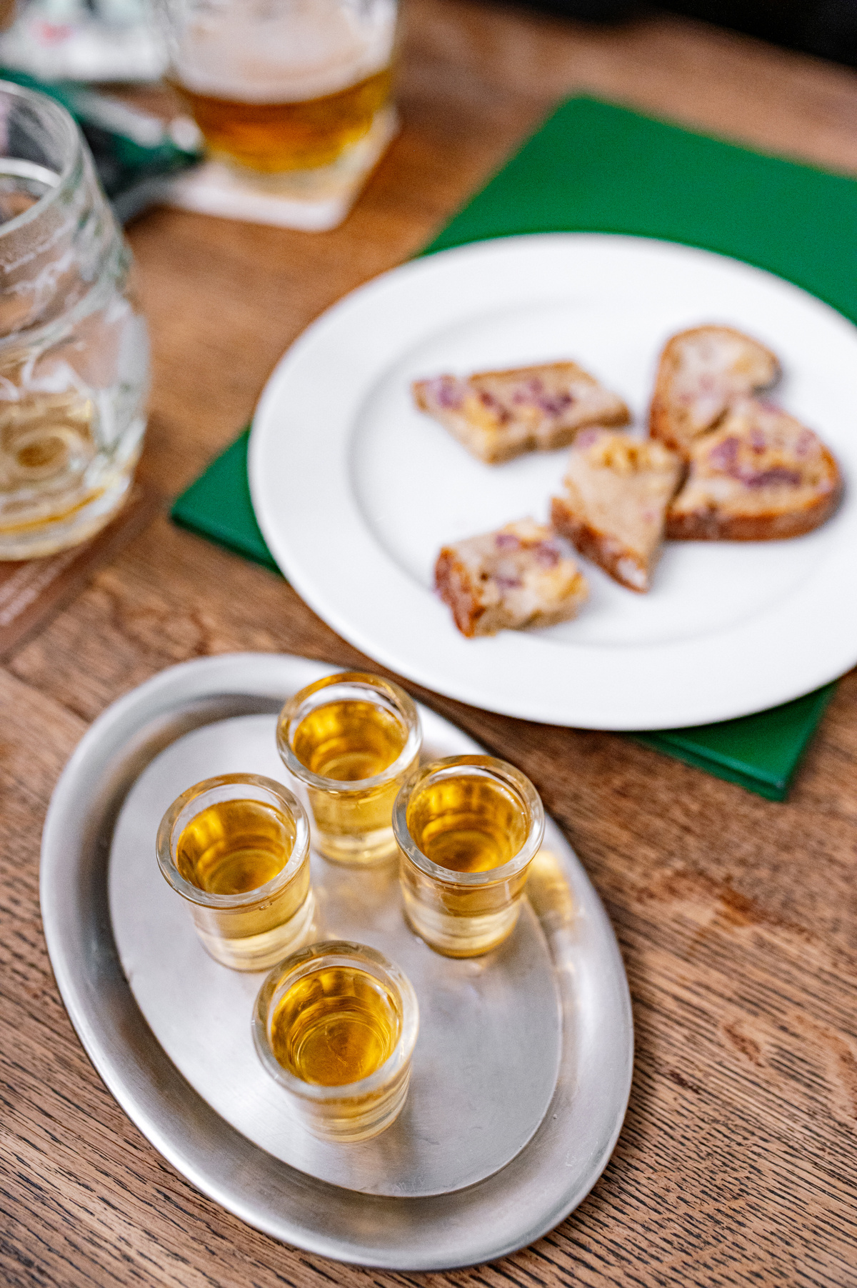 Čtyři panáky zlatavého alkoholu na kovovém podnose a talíř s kousky chleba s pomazánkou na dřevěném stole.