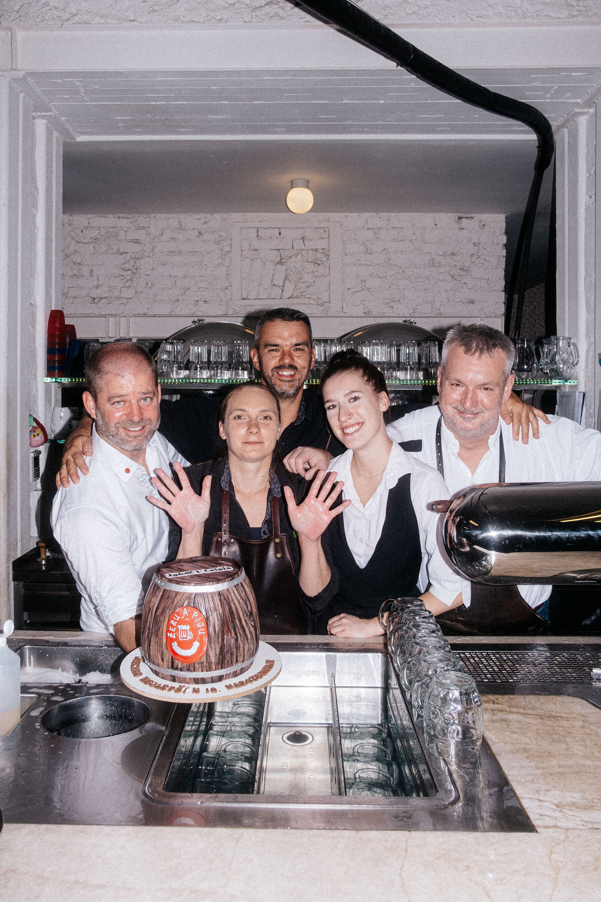 Tým restaurace pózuje společně za barem vedle dortu ve tvaru pivního sudu při oslavě výročí.