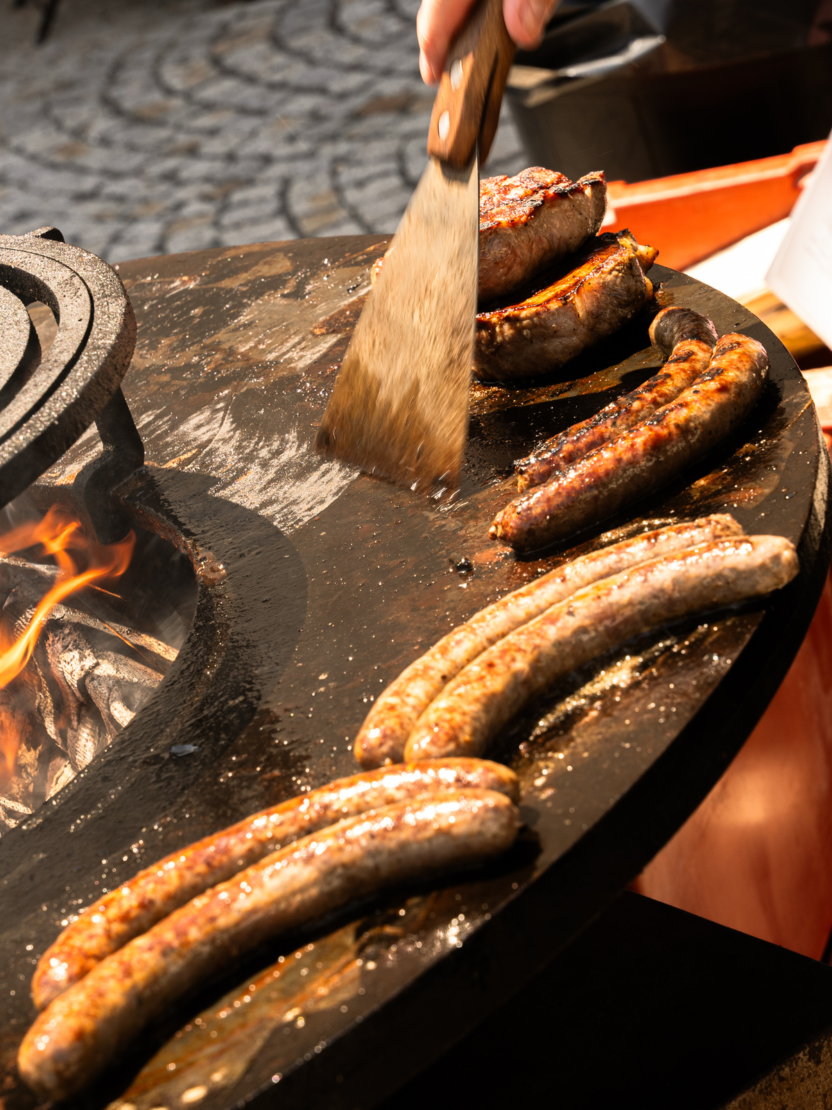 Klobásy a steaky se opékají na venkovním grilu nad otevřeným ohněm.