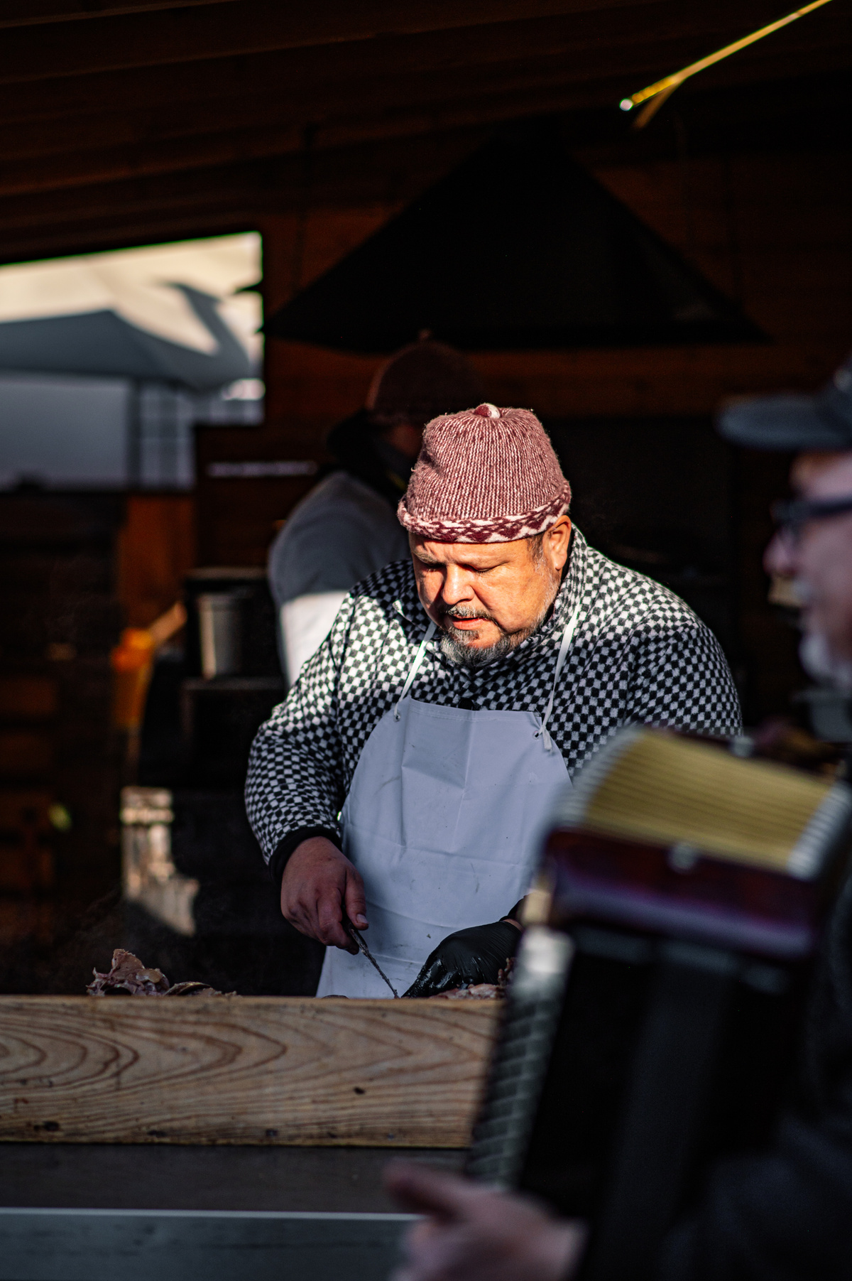 Muž v zástěře a pletené čepici krájí maso při venkovní zabijačce, v popředí je vidět akordeonista.
