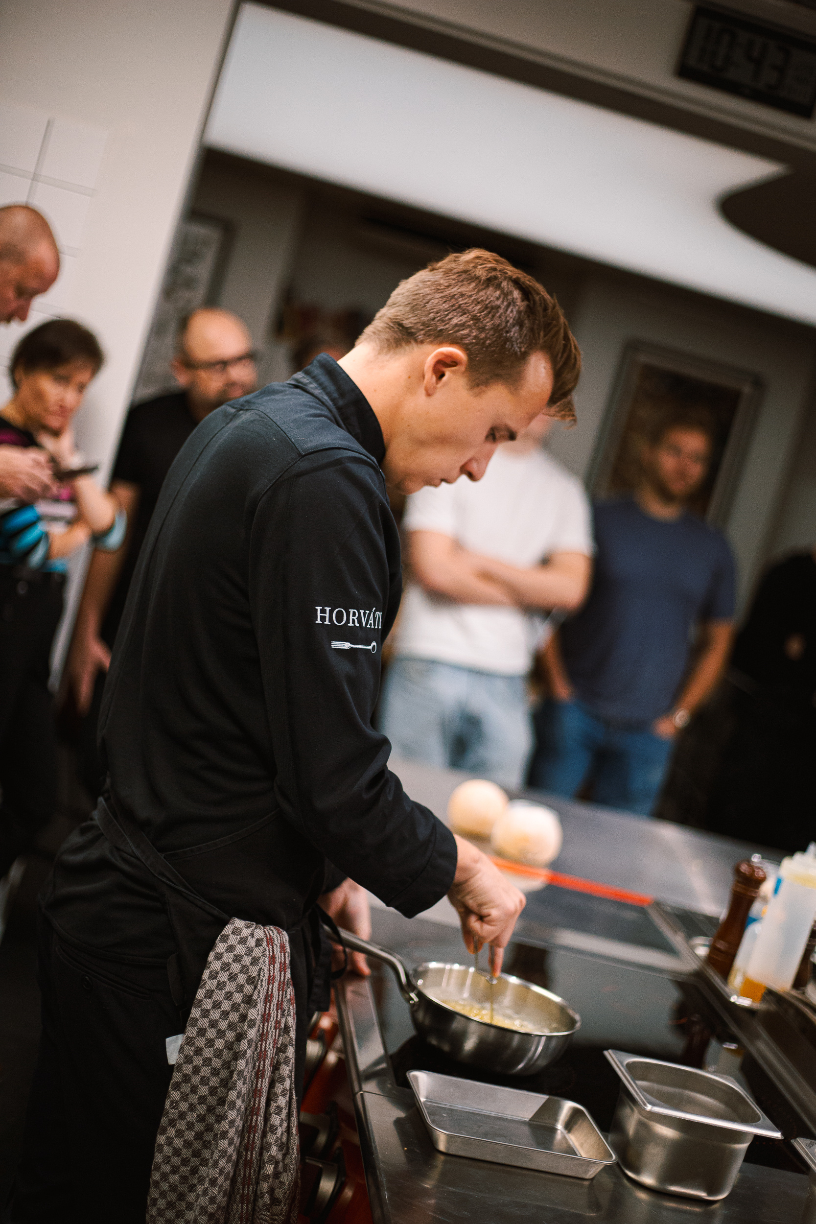 Kuchař v černé uniformě připravuje jídlo na pánvi při kuchařské ukázce, za ním sledují lidé.