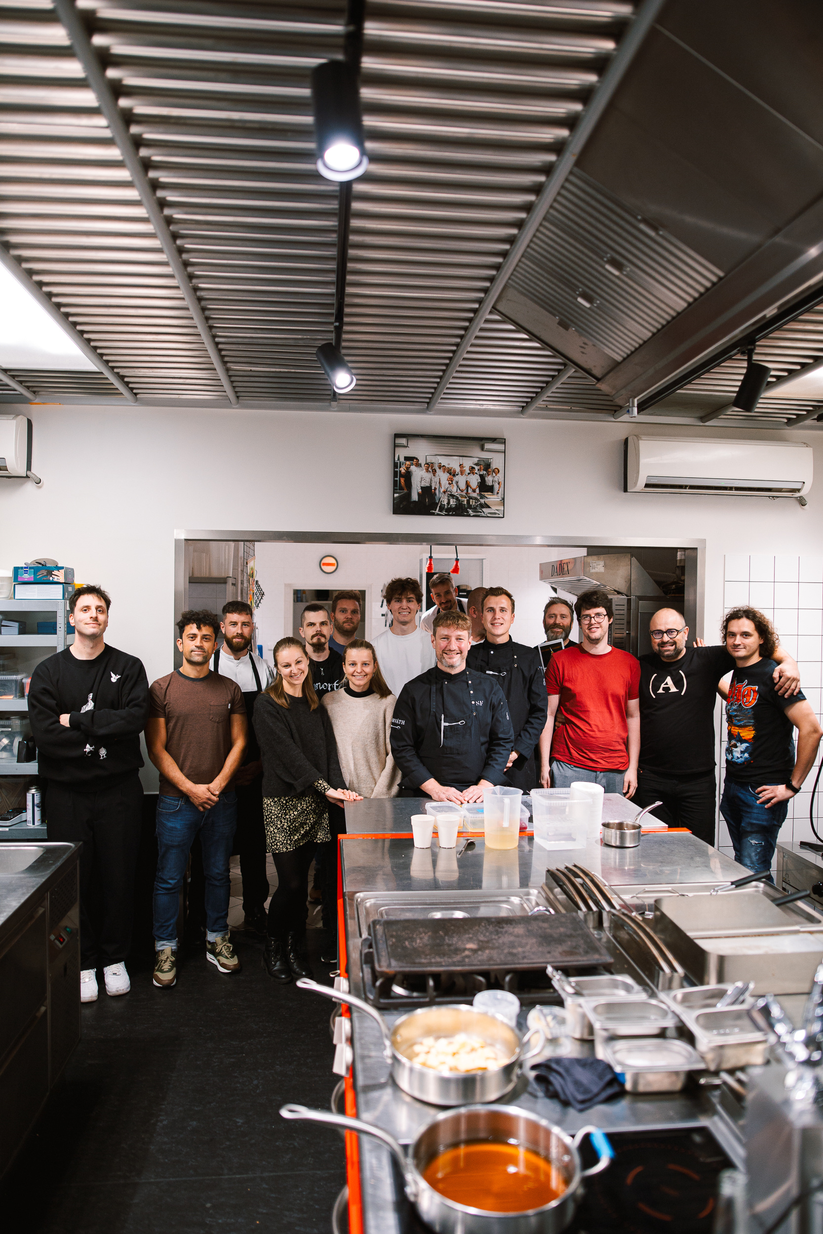 Skupina lidí stojící v profesionální kuchyni, kuchaři a účastníci kurzu pózují u pracovního stolu s nádobím a pánvemi.