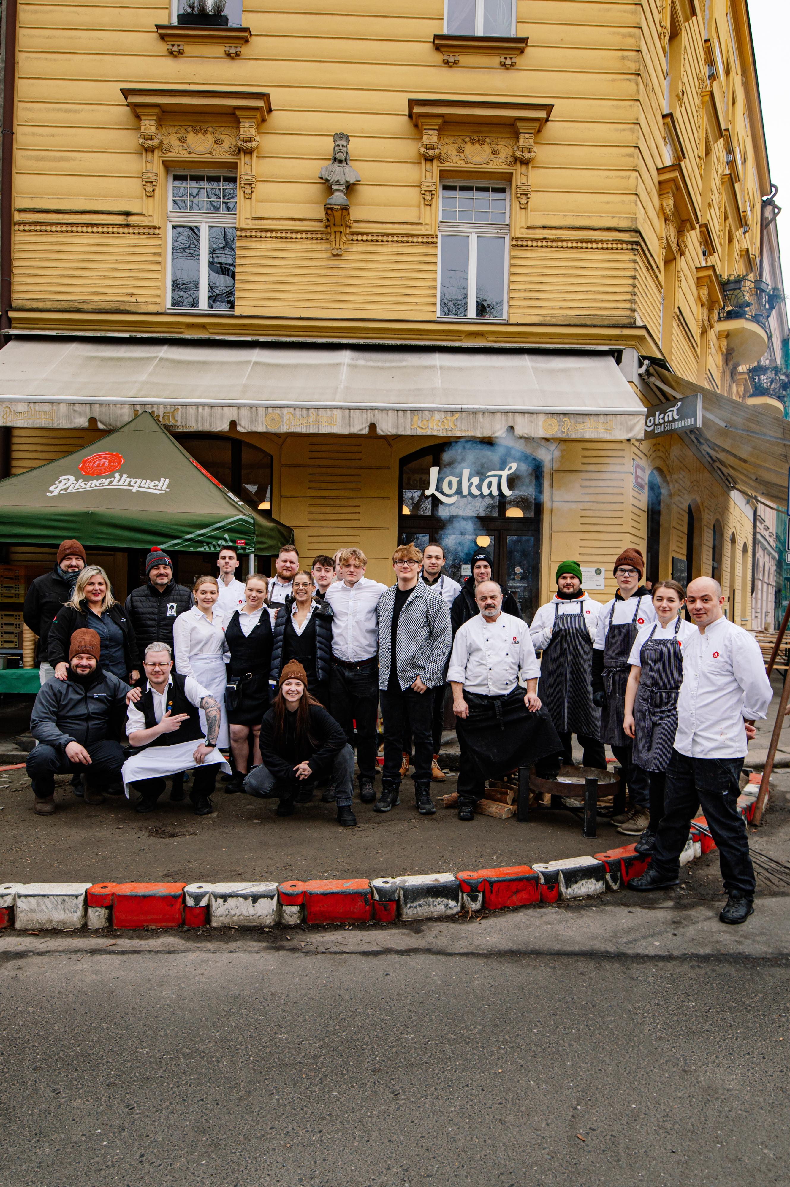 Skupinová fotografie personálu podniku Lokál Nad Stromovkoupřed budovou, kuchaři a obsluha pózují na ulici před podnikem.