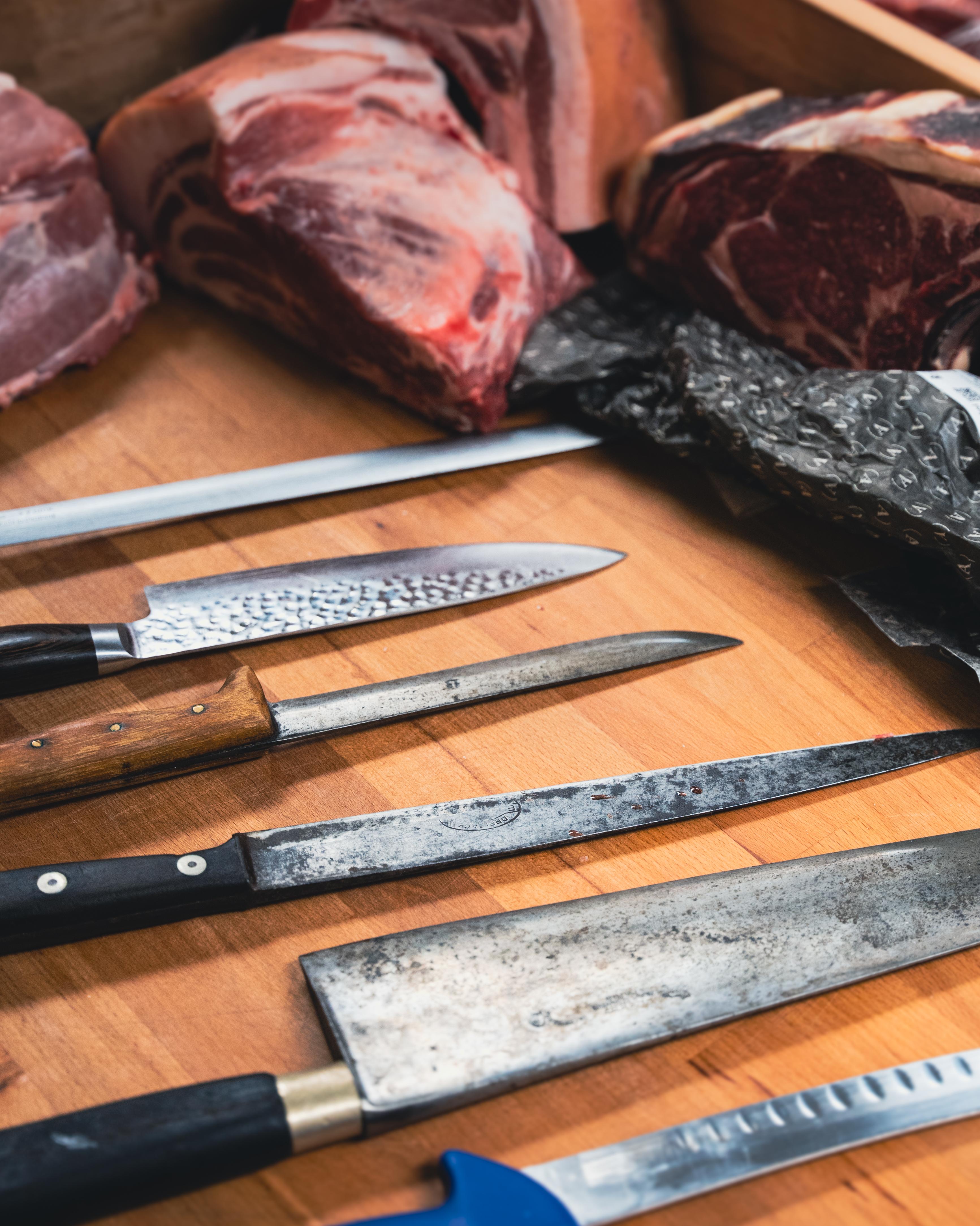 Řeznické nože na dřevěném stole vedle kusů syrového masa.