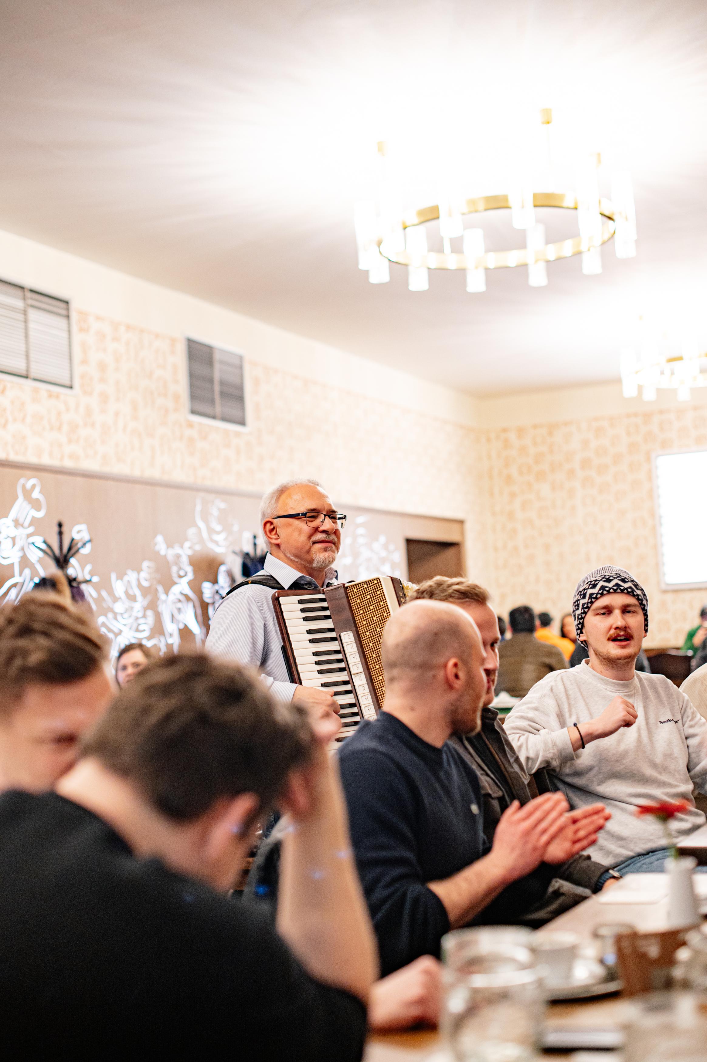 Muž hrající na akordeon mezi hosty v restauraci, lidé u stolu zpívají a tleskají.