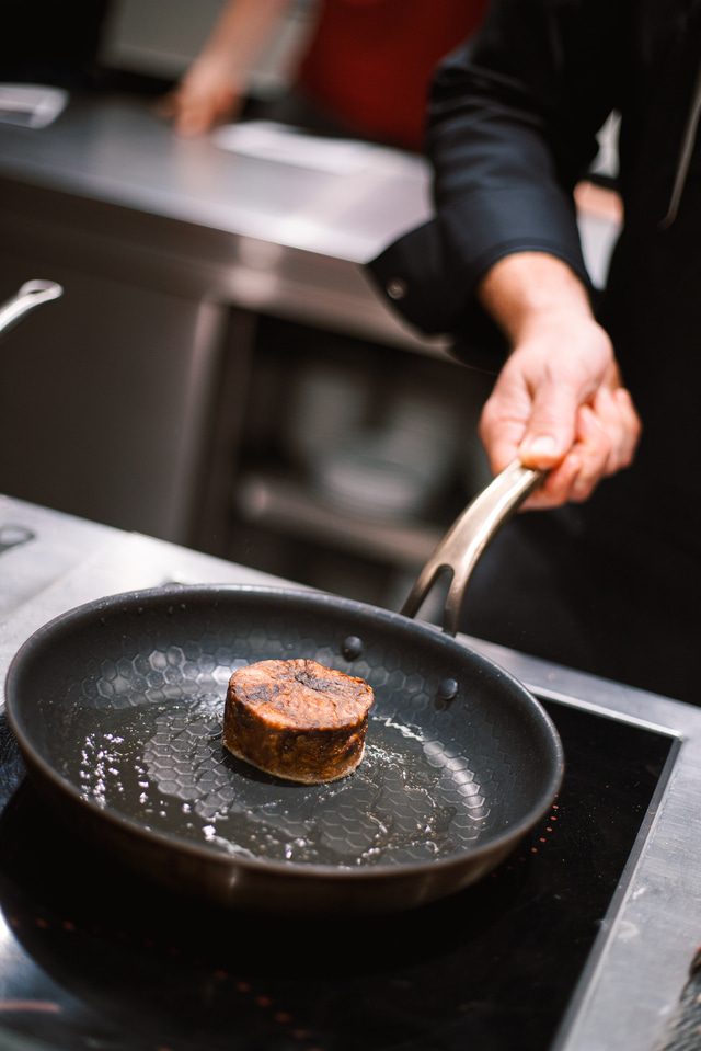 Kuchař držící pánev s opečeným kusem masa při přípravě pokrmu v profesionální kuchyni.